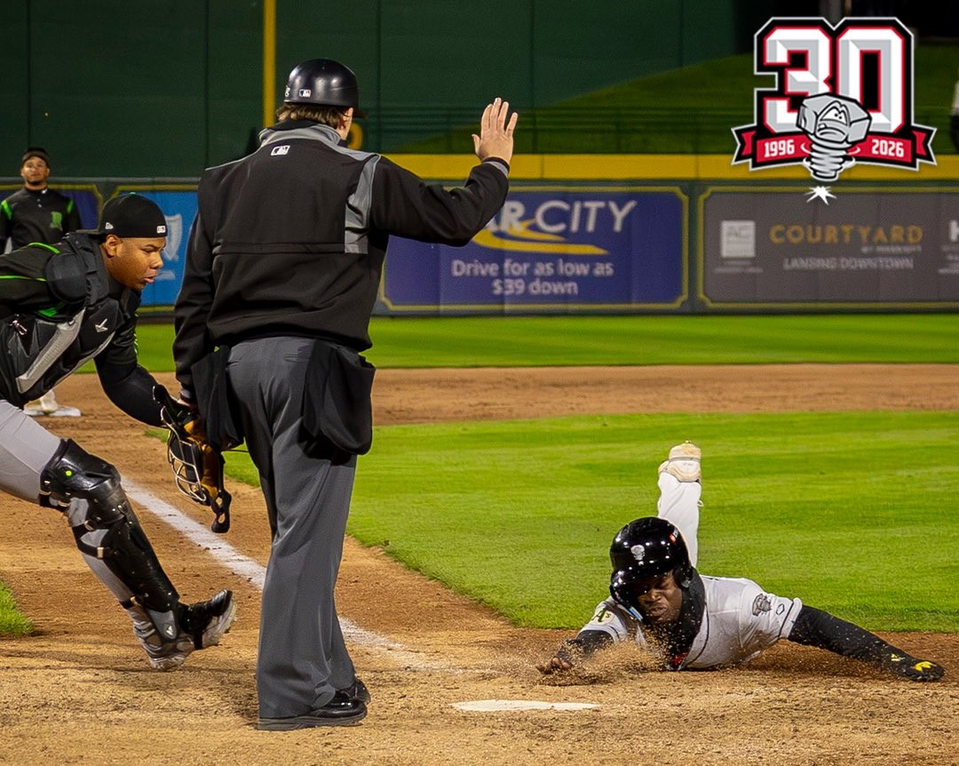 Baseball play at home plate: a sliding runner reaches the plate as the catcher and umpire react, with a 30th anniversary logo in the top-right.