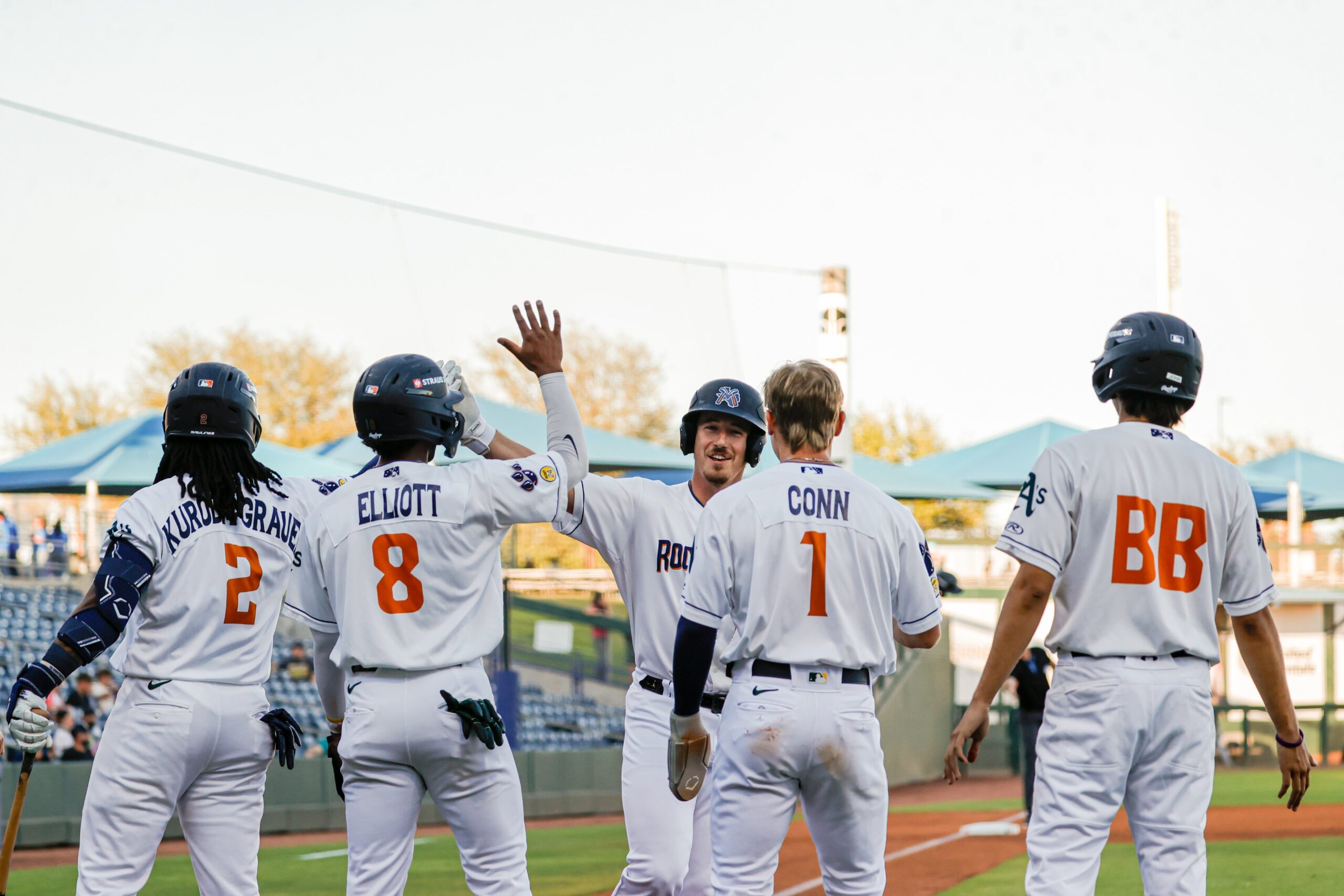 Baseball players in white uniforms celebrate with a high-five on the field near the dugout.