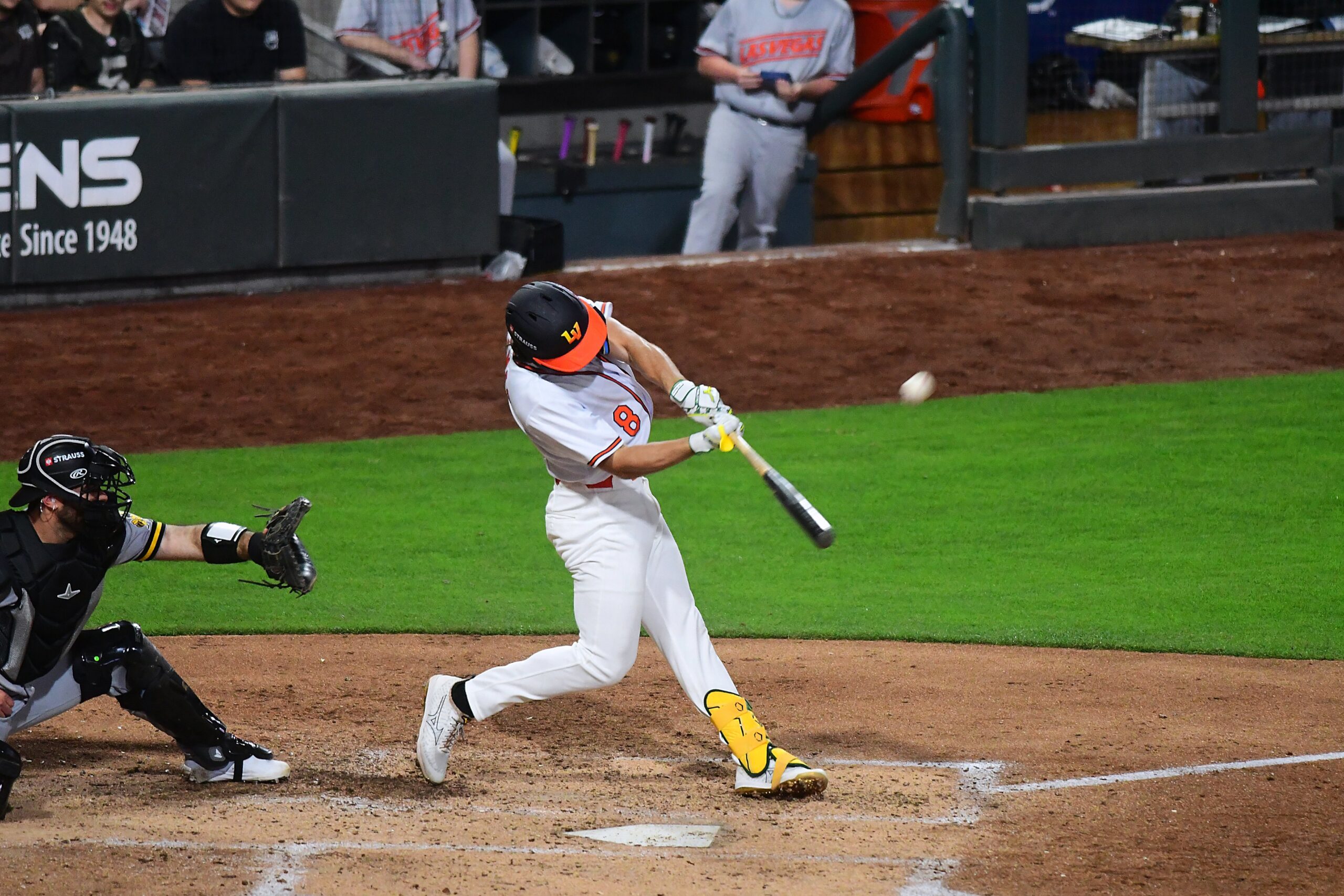 Baseball batter in white uniform swinging at a pitch near home plate, ball just off the bat, catcher diving in black gear nearby.