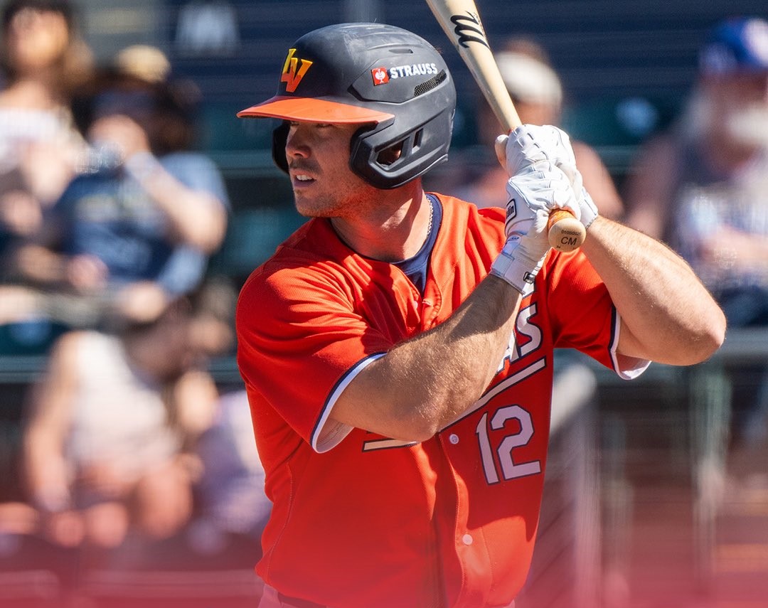 Baseball player in red jersey swinging a bat, wearing a black helmet with the number 12 visible and white gloves; crowd in the background.