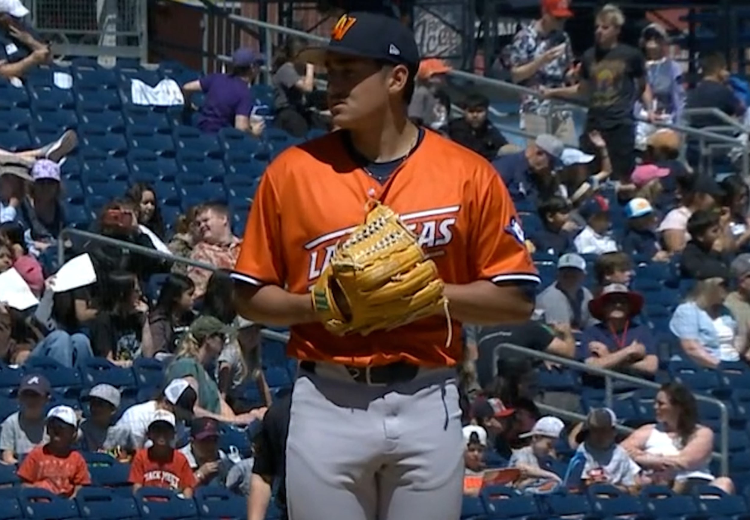 Baseball player in orange uniform holding a glove, standing on the field with spectators in the background.