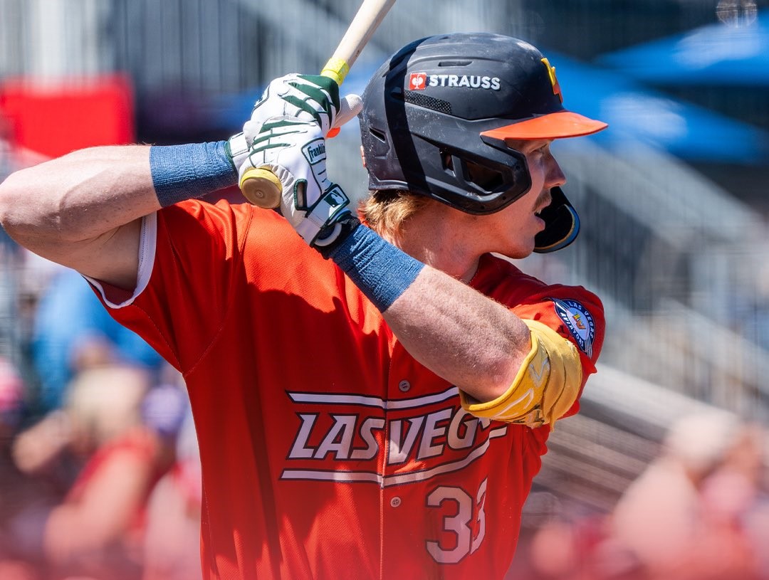 Baseball player in a red Las Vegas jersey swings a bat during a game, wearing a black helmet and blue wristbands with a yellow guard.