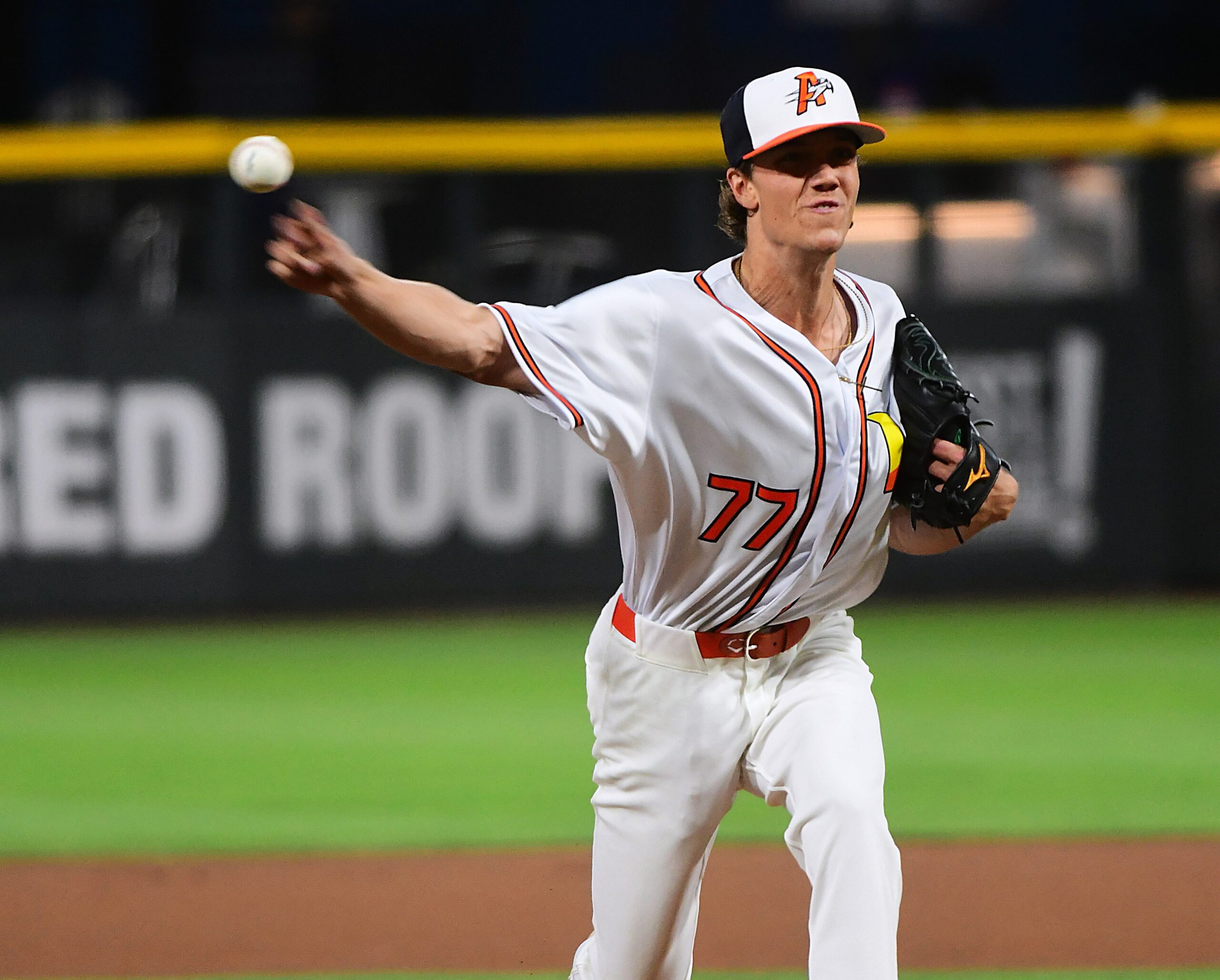 Baseball pitcher in a white uniform throwing a pitch on the mound, left arm extended and ball in motion behind him (jersey number 77).