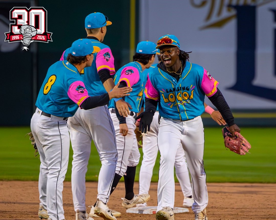 Baseball players in turquoise and pink uniforms celebrate with a group high-five on the infield.