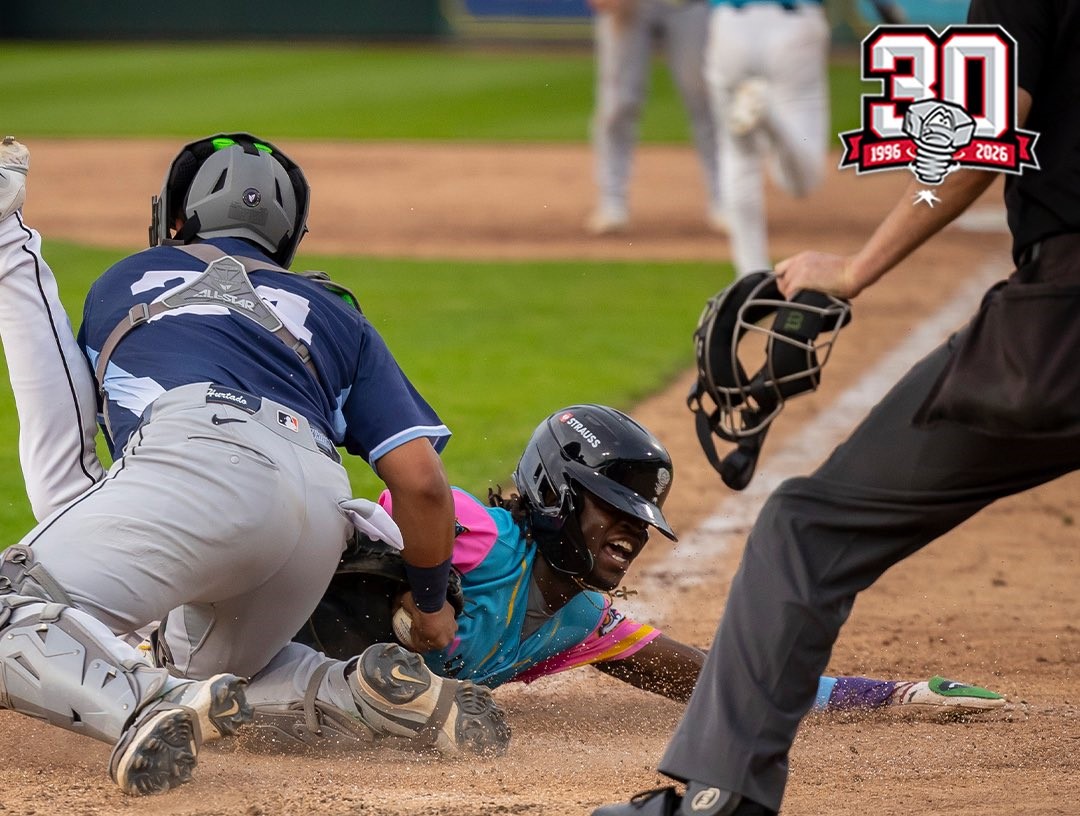Baserunner in teal slides headfirst into home plate as the catcher in gray gear reaches to tag him; umpire nearby with mask in hand and dust rising.