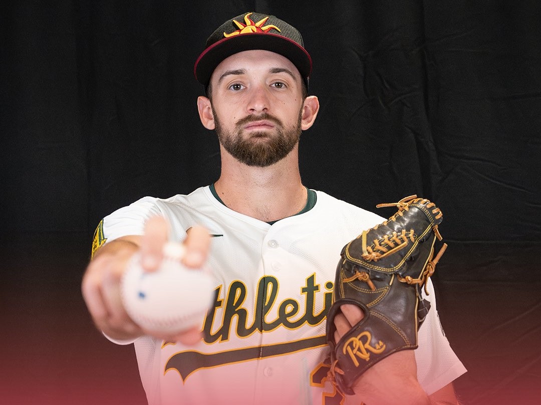 Baseball pitcher in a white Athletics jersey extends his arm toward the camera, gripping a baseball with a brown glove on the other hand, wearing a dark cap with a yellow logo.