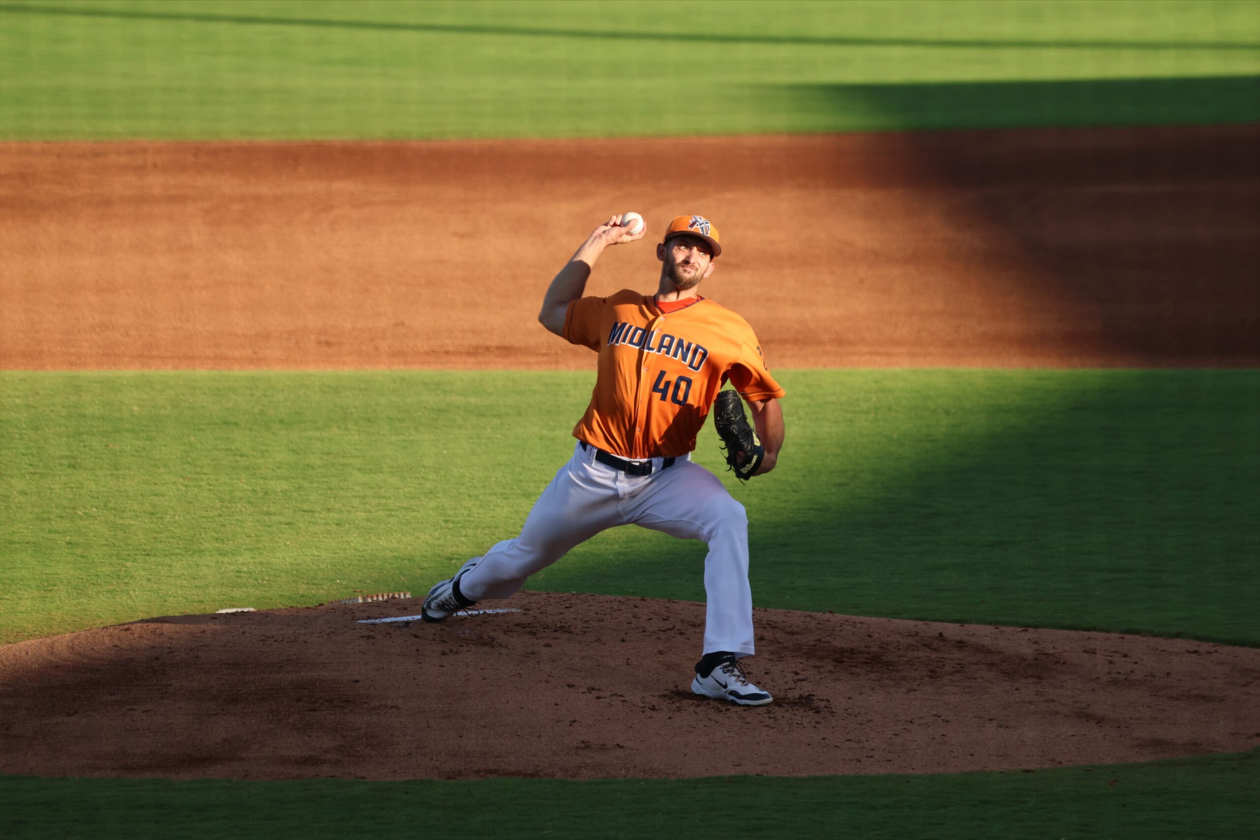 Baseball pitcher in orange Midland uniform delivering a pitch from the mound on a green field.