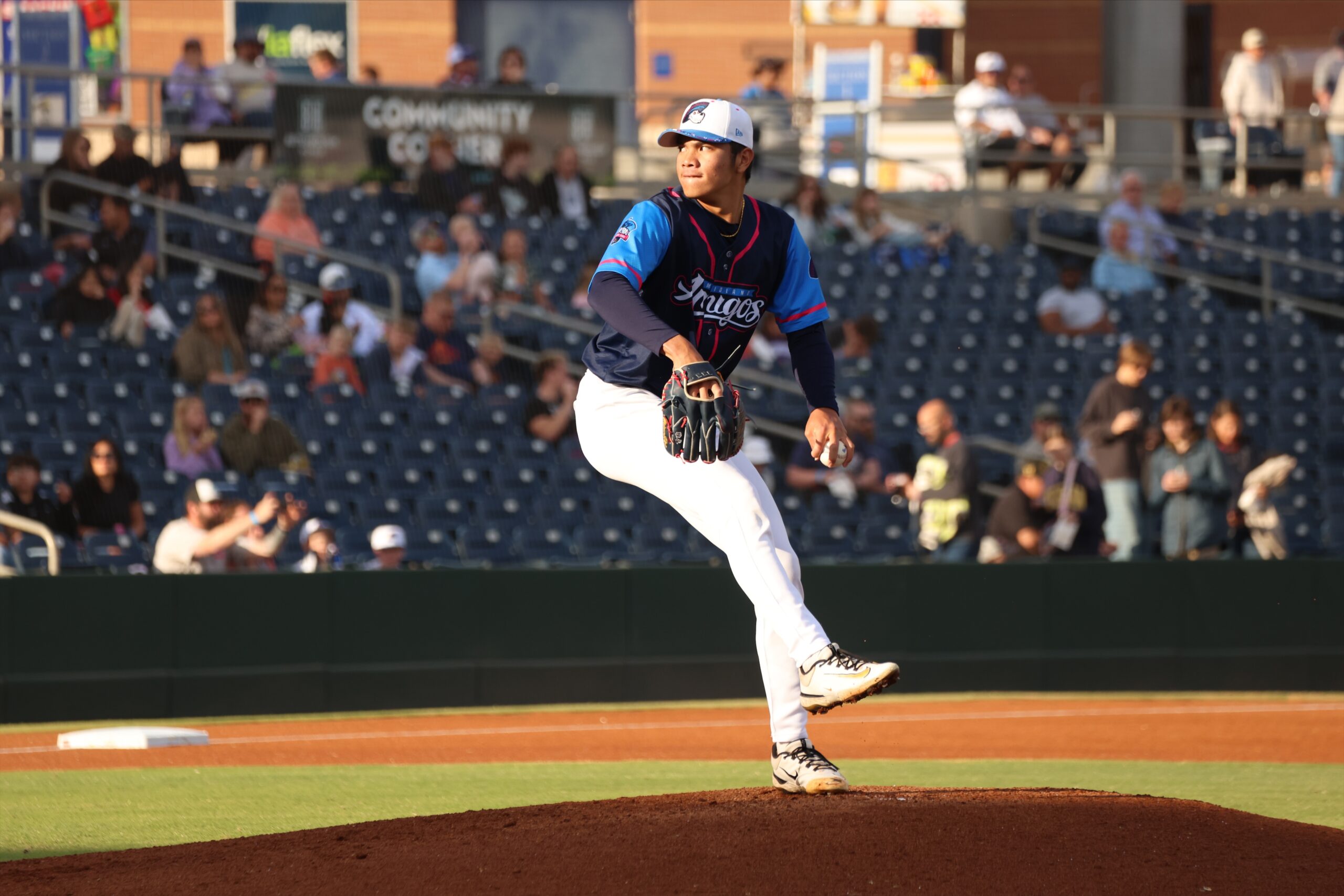 Baseball pitcher in blue and navy uniform throwing a pitch on the mound with a blurred crowd in the stands behind him.