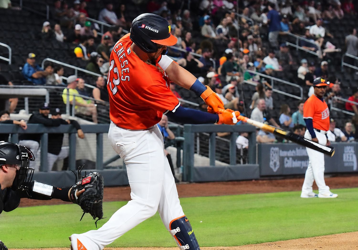 Baseball batter in an orange uniform swings a bat during a game with a catcher ready behind him.