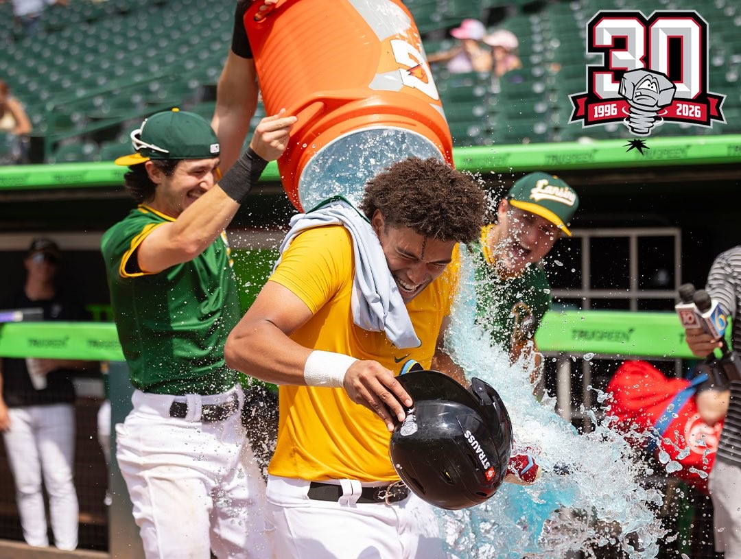 Baseball player in a yellow jersey gets drenched with a large orange cooler as teammates celebrate nearby at a stadium, with a 30th anniversary logo visible in the top-right corner.