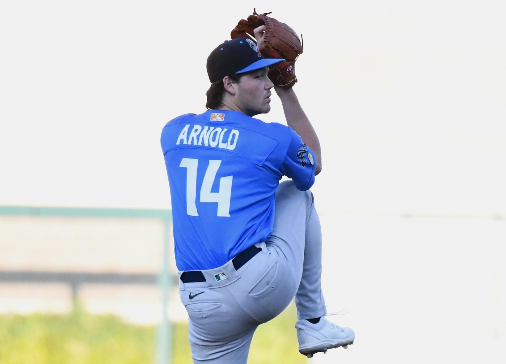 Baseball pitcher in a blue jersey (Arnold, No. 14) mid-throw with glove raised above his head in a stadium setting