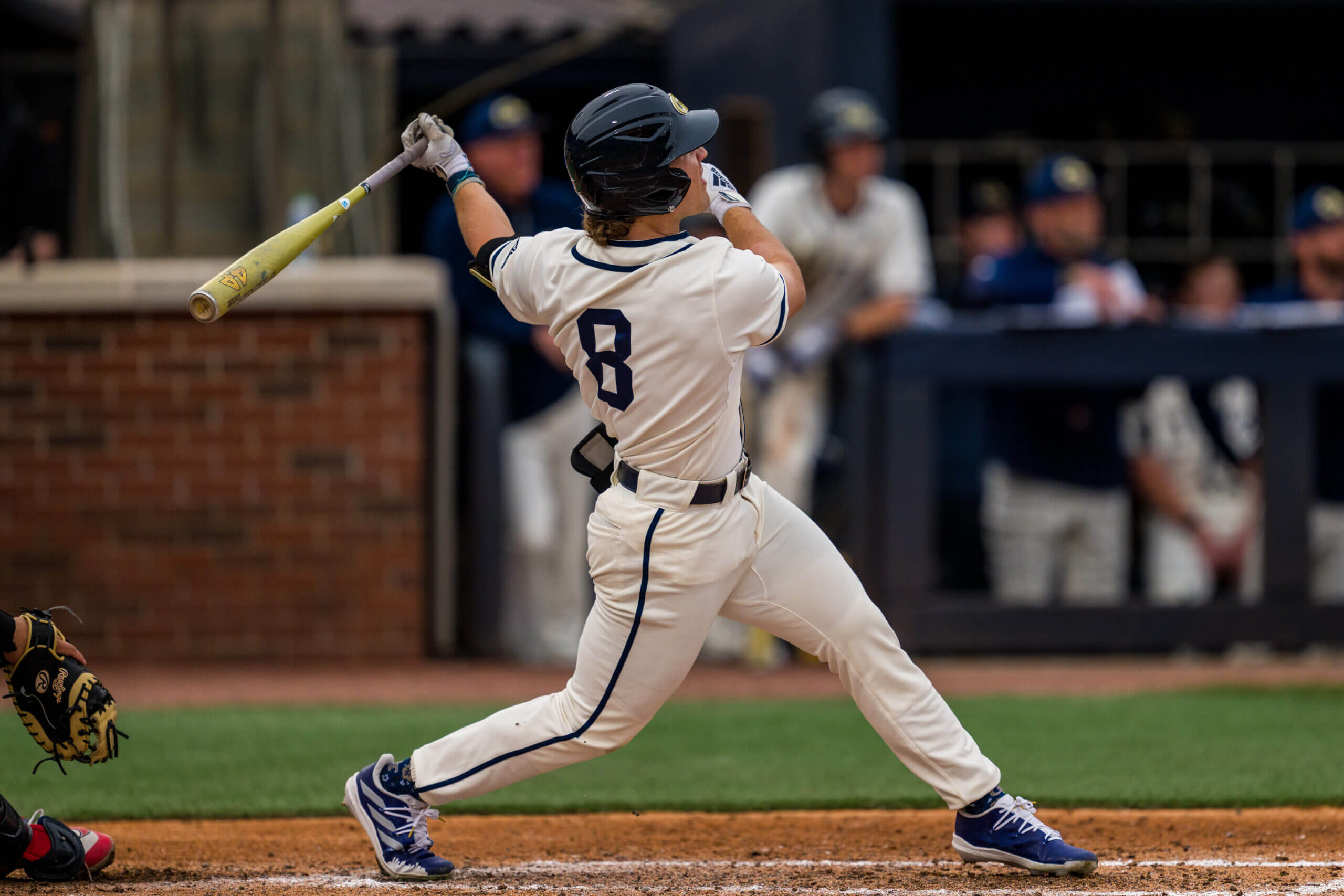 Baseball batter in a white uniform swinging the bat during a game, helmet on, in the batter's box with a glove nearby.