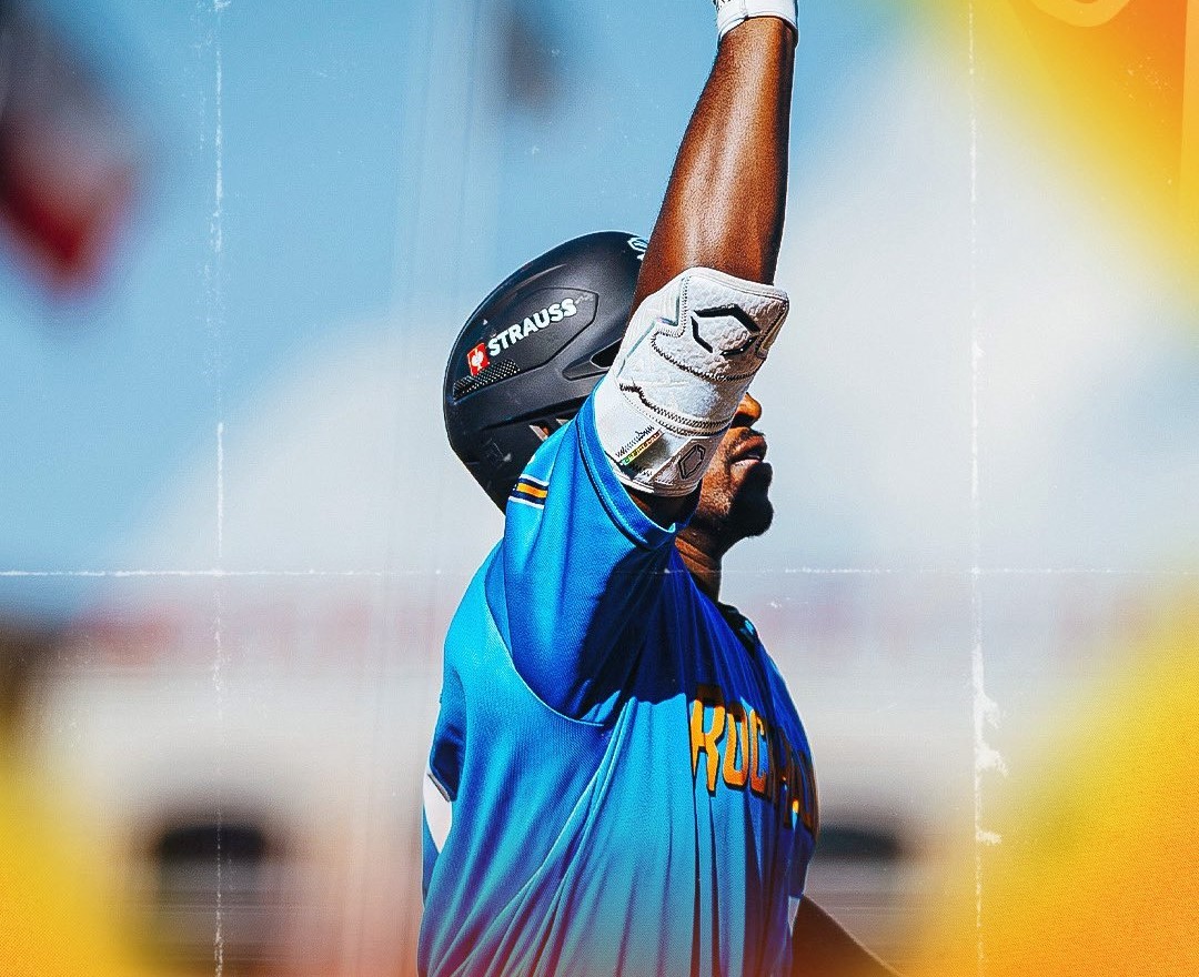 Cricketer in a blue jersey raises his bat and helmet in celebration on the field, orange sunlight in the background.