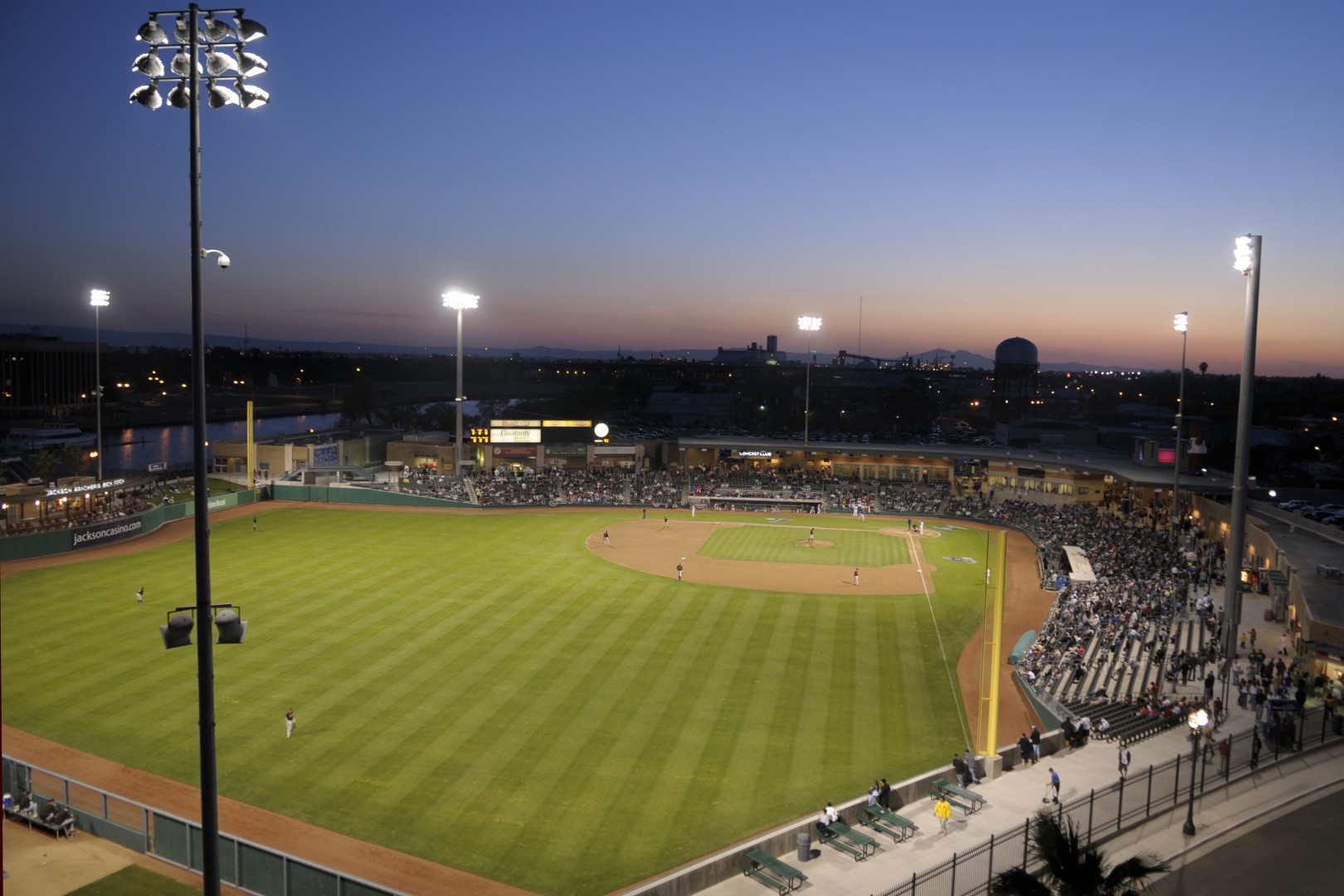 Wide view of a baseball stadium at dusk, with players on the field and bright stadium lights over the stands packed with fans.