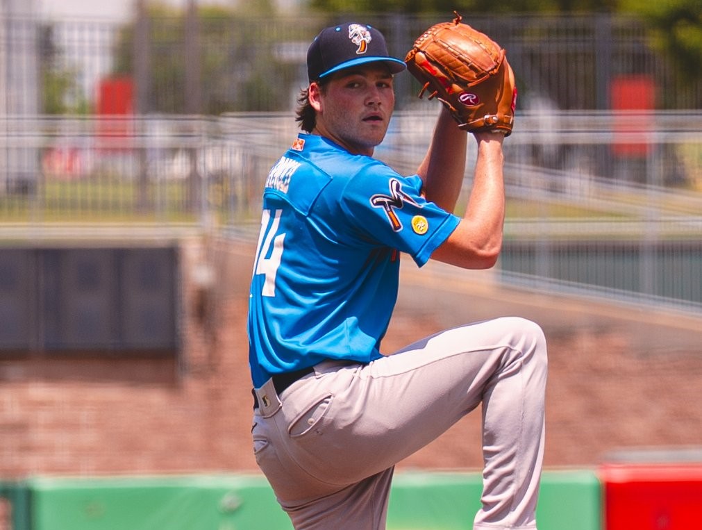 Baseball pitcher in a blue jersey throws a pitch on the mound with his leg raised.
