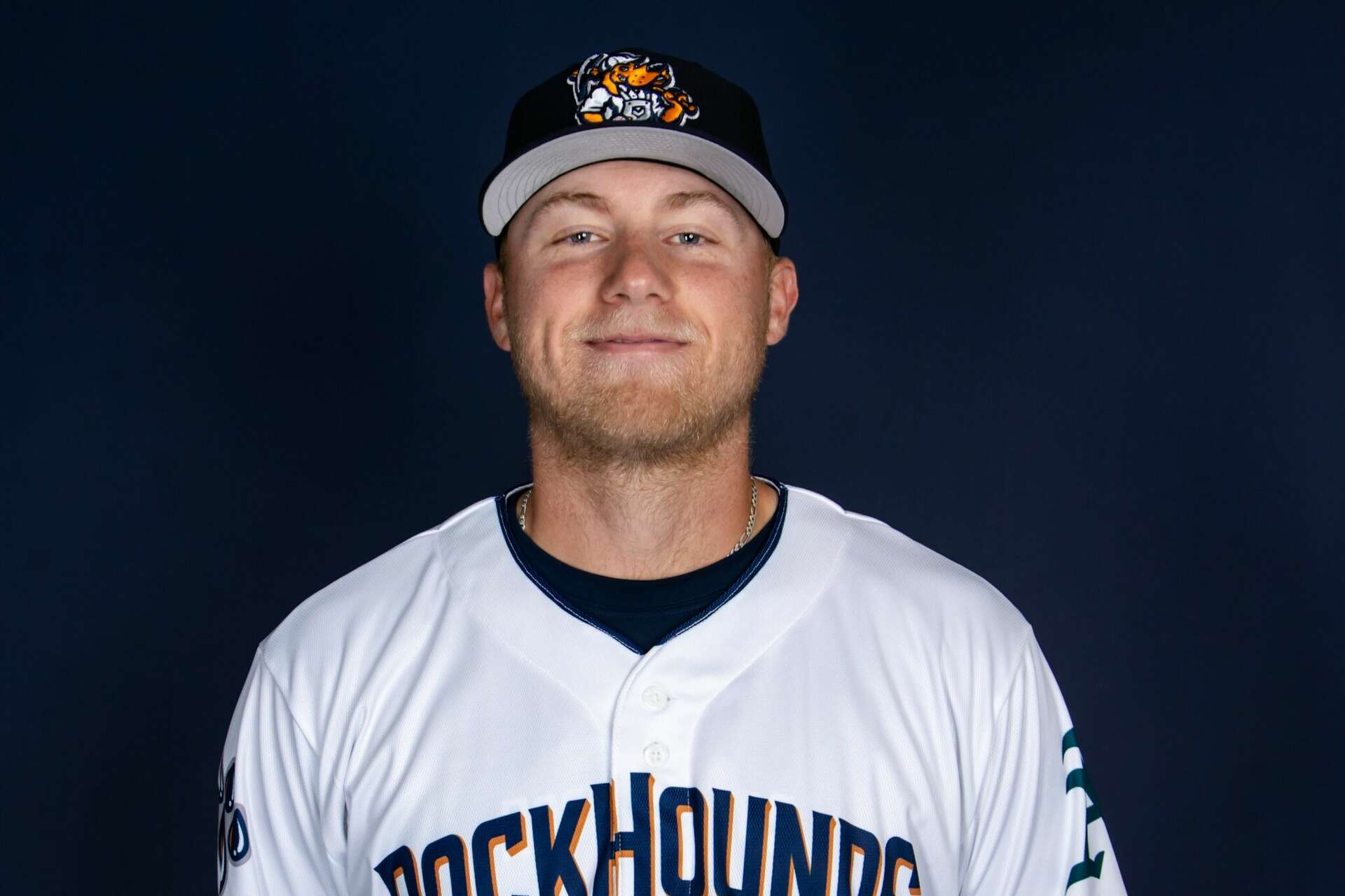 Portrait of a baseball player wearing a white RockHounds jersey and a black cap with a colorful logo, posed against a dark blue background.