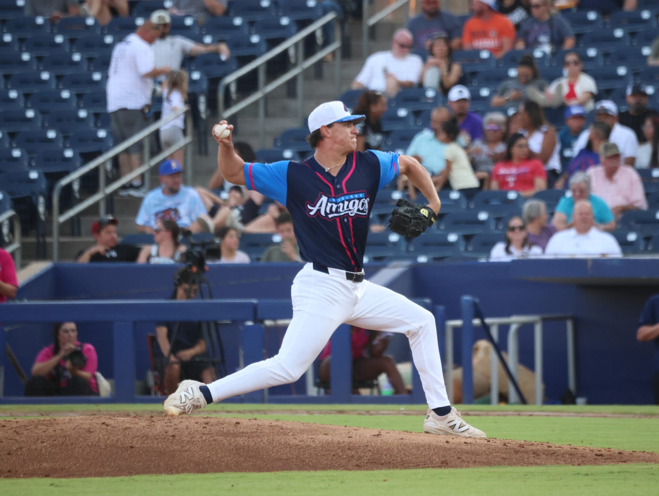 Baseball pitcher in a blue and navy uniform with a white cap mid-pitch on the mound, crowd blurred in the stands behind him.