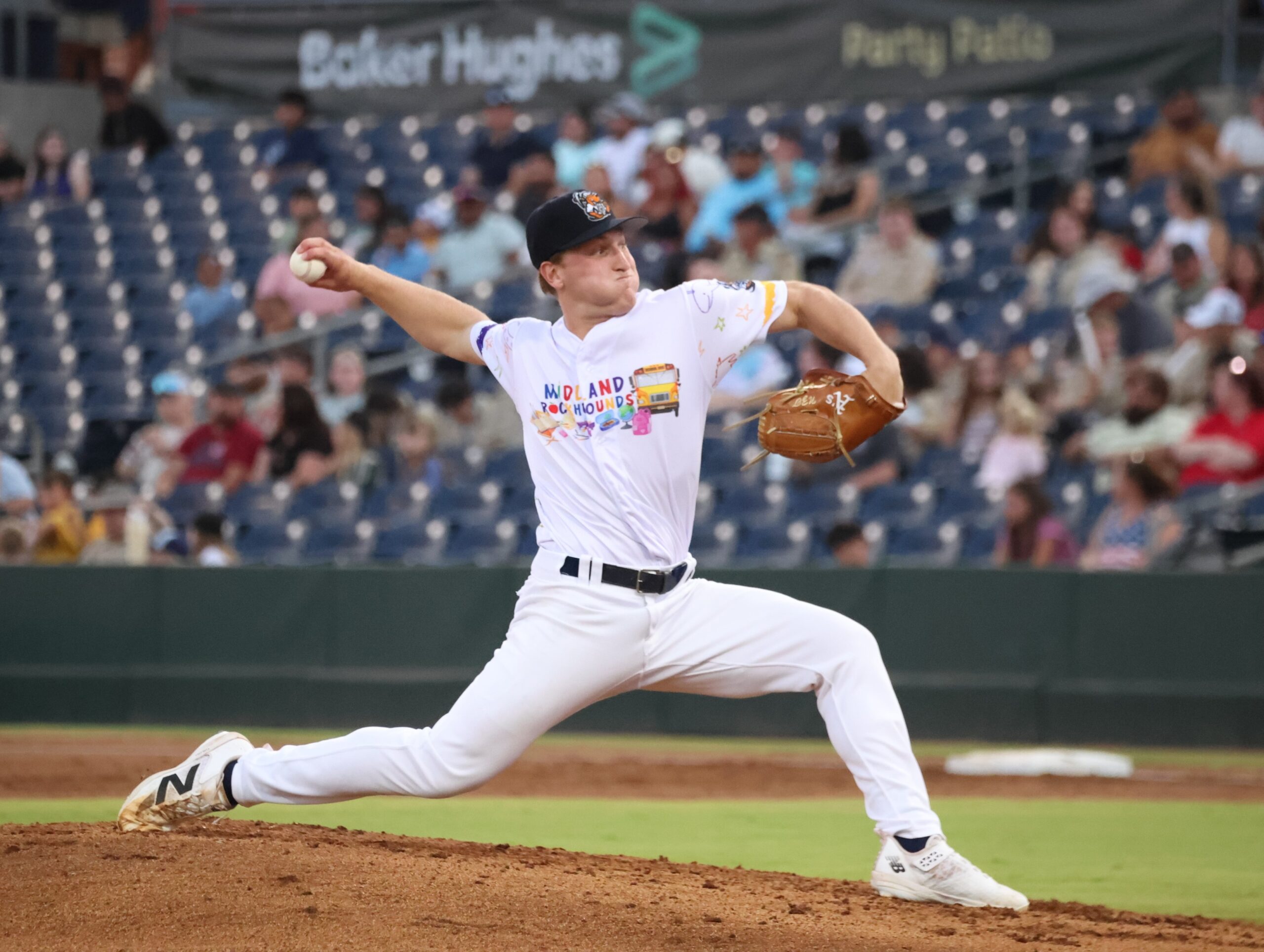 Baseball pitcher in a white Midland RockHounds uniform delivering a pitch from the mound, with a brown glove on his right hand and crowd in the stands behind.